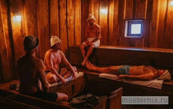 Мужчина в банной шапке сидит в предбаннике с чашкой чая, на заднем плане виден мягкий свет из открытой парной.